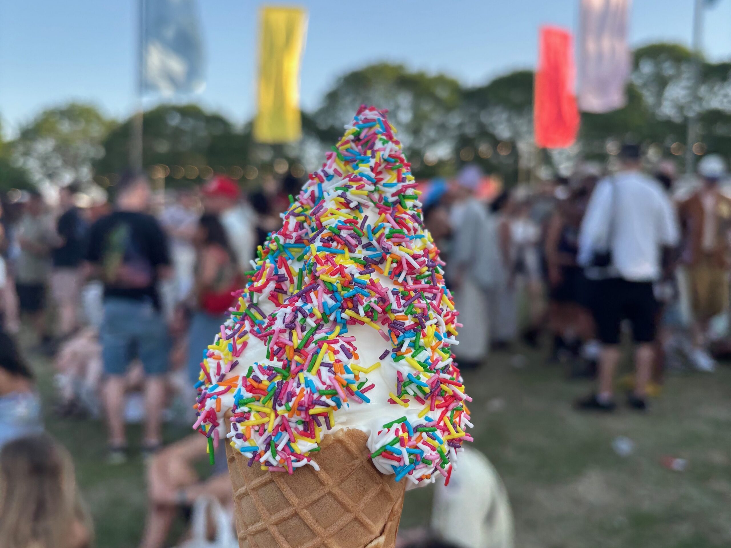 A ice cream cone covered in hundreds-and-thousands in the foreground, a festival in the background